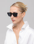 A woman with blonde hair pulled back wears For Art's Sake® Harmony(S) black minimal sunglasses and a white satin shirt with a popped collar, posing against a plain light gray background.