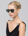 A woman with blonde hair in a neat bun wears For Art’s Sake® Harmony(S) sculptural rectangular sunglasses and a black sleeveless top with gold shoulder buttons, set against a plain light gray background.