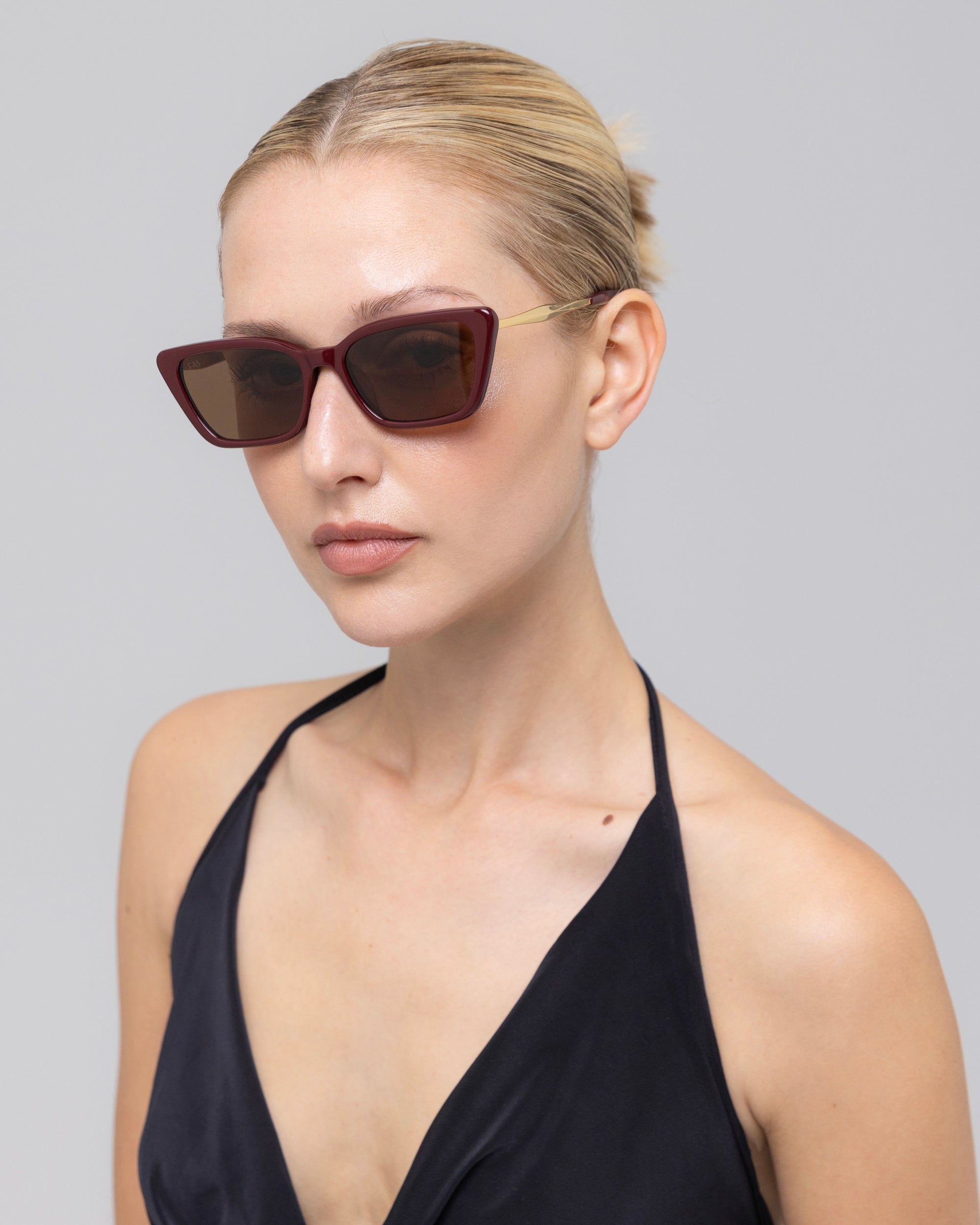 A woman with blond hair in a sleek bun wears For Art's Sake® Harmony(S) sculptural cat-eye sunglasses with dark red frames and a black halter top, posing against a plain light gray background.