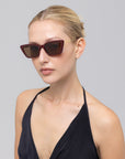 A woman with blond hair in a sleek bun wears For Art's Sake® Harmony(S) sculptural cat-eye sunglasses with dark red frames and a black halter top, posing against a plain light gray background.