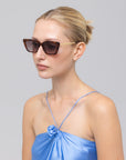 A woman with blonde hair in a bun wears For Art's Sake® Harmony(S) boxy sunglasses and a shiny light blue spaghetti strap top with a flower detail, posing against a plain light gray background.