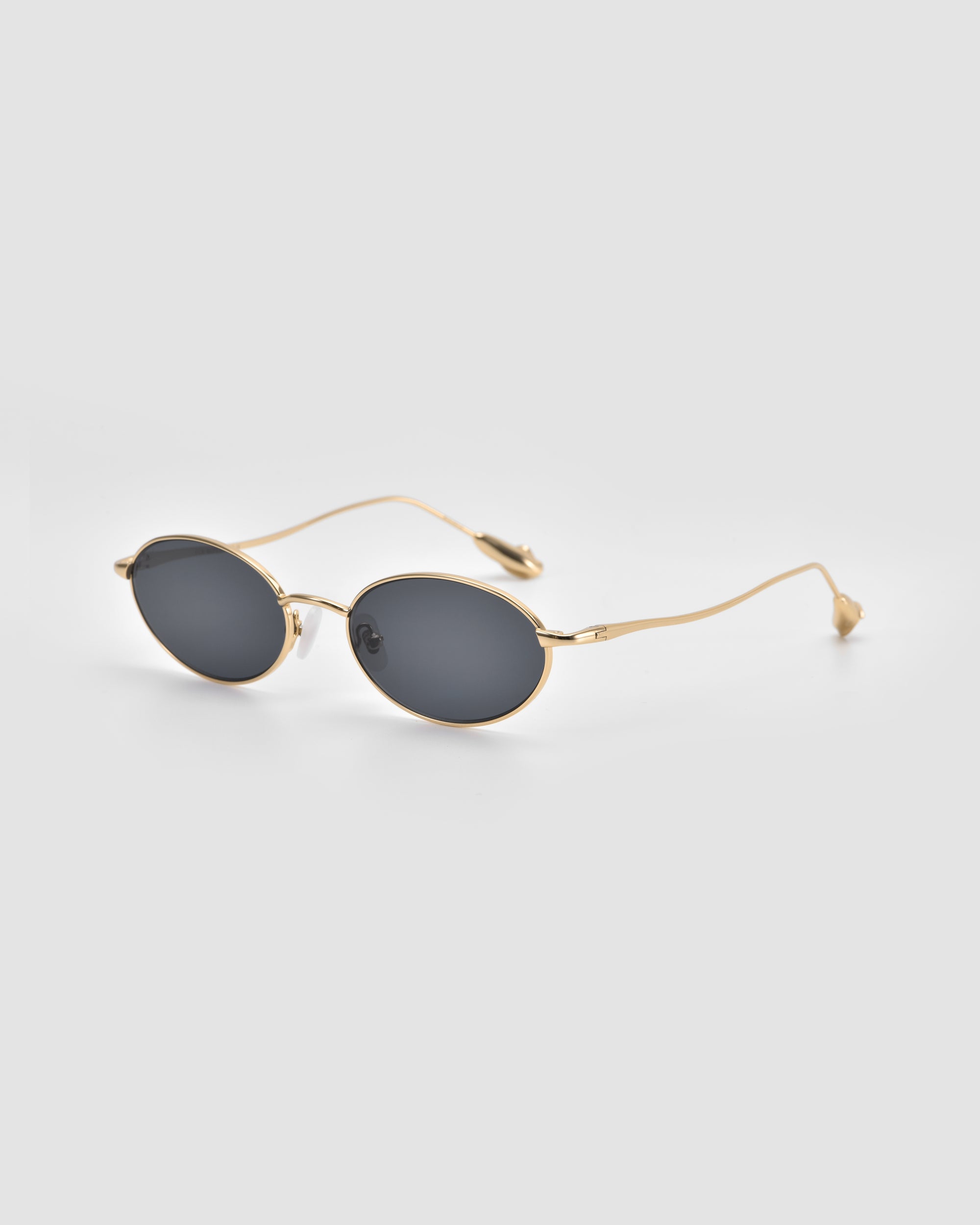 The Calla I by For Art's Sake® are oval-shaped sunglasses with dark lenses and thin gold metal frames. Shown on a light gray background, the image emphasizes their modern, lightweight sculptural design.