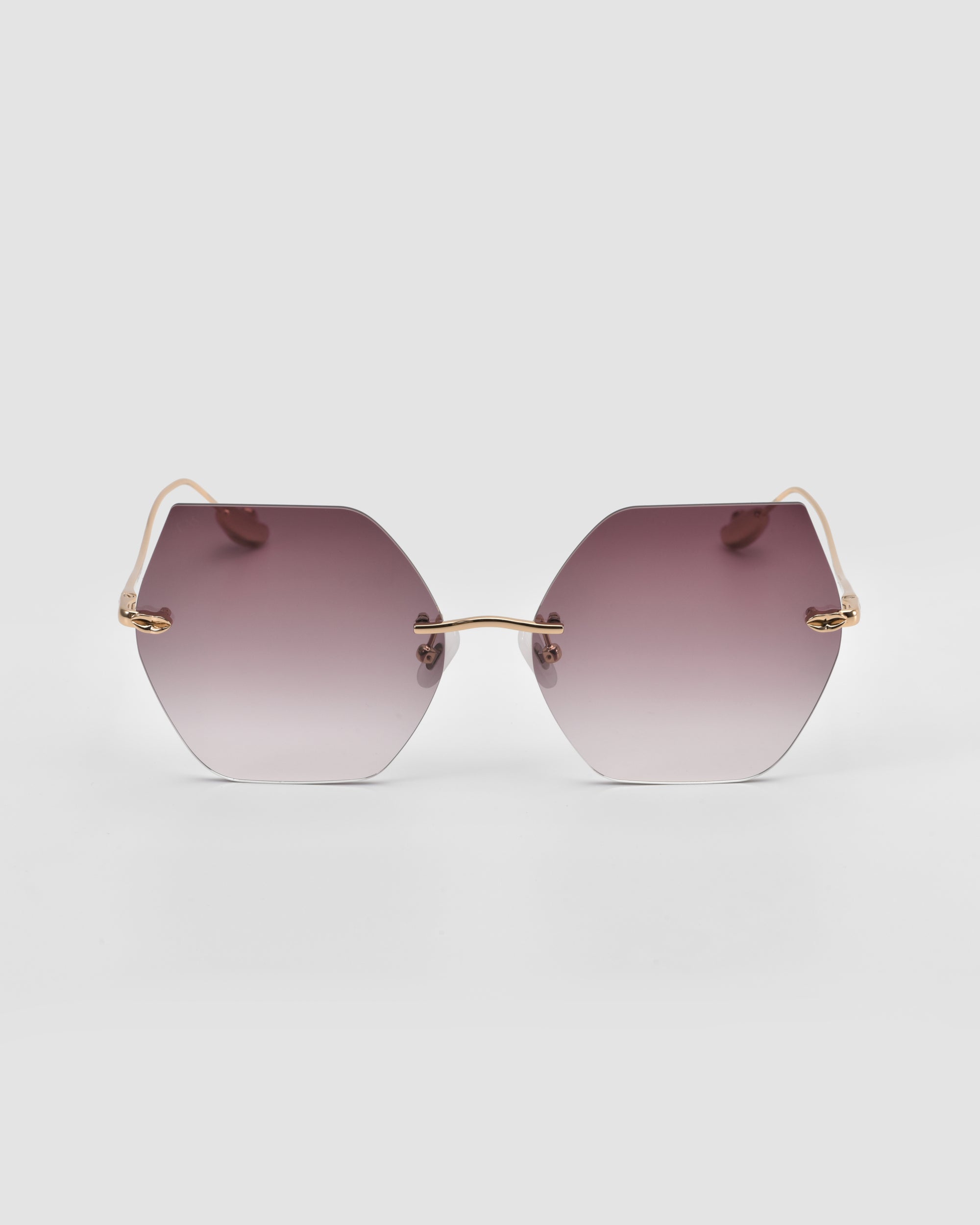 The Georgia by For Art’s Sake® features hexagonal oversized frameless sunglasses with gold wire arms and gradient purple lenses. Sculpted metal temples elevate this modern accessory, shown against a plain light gray background.