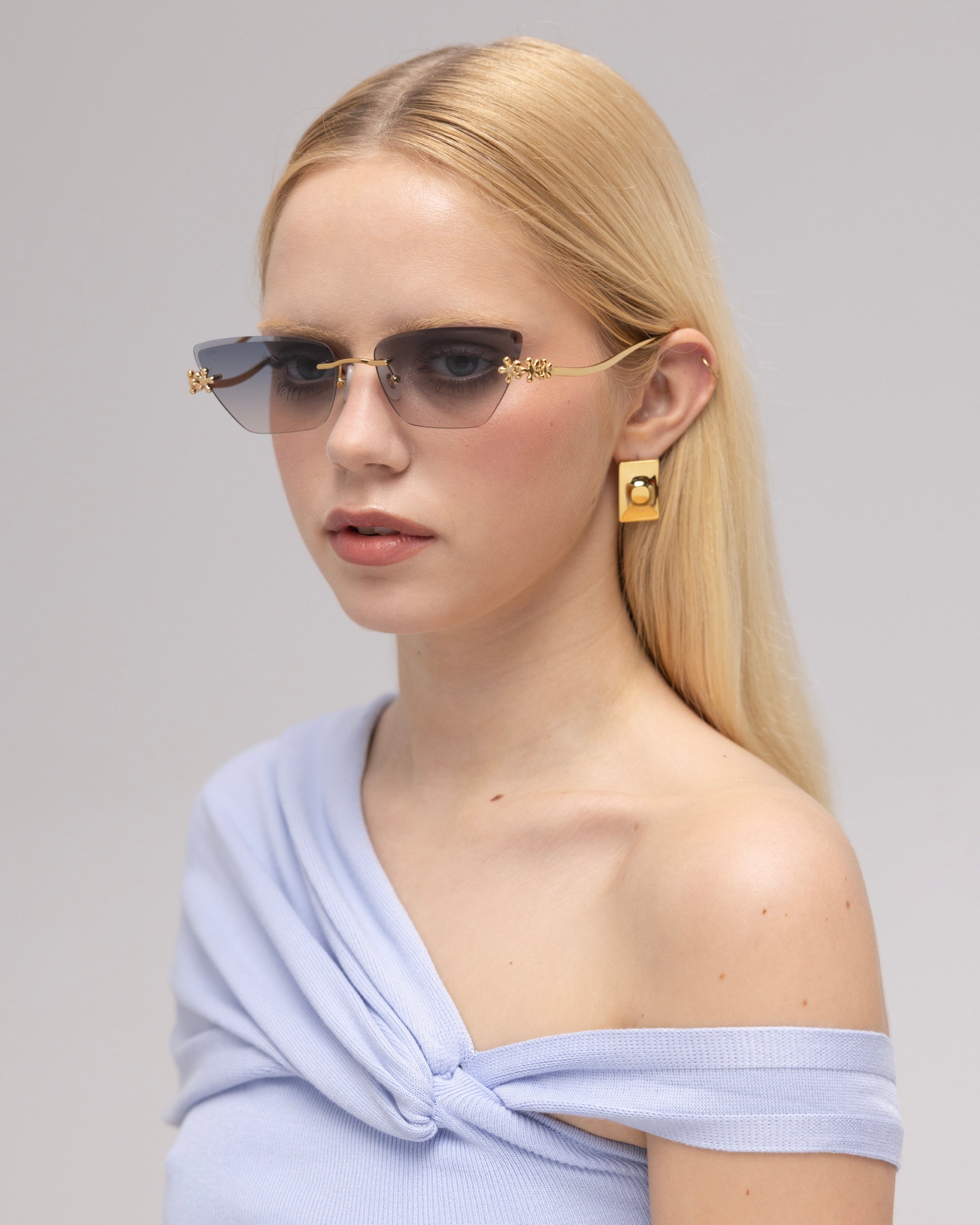 A woman with straight blonde hair wears Florist blue-tinted frameless sunglasses by For Art's Sake®, gold geometric earrings, and an off-the-shoulder light blue top, posing against a neutral background.