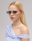 A woman with straight blonde hair wears Florist blue-tinted frameless sunglasses by For Art's Sake®, gold geometric earrings, and an off-the-shoulder light blue top, posing against a neutral background.