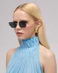 A woman with long blonde hair wears the Florist black rectangular sunglasses by For Art's Sake®, gold geometric earrings, and a sleeveless light blue pleated high-neck top against a plain background.