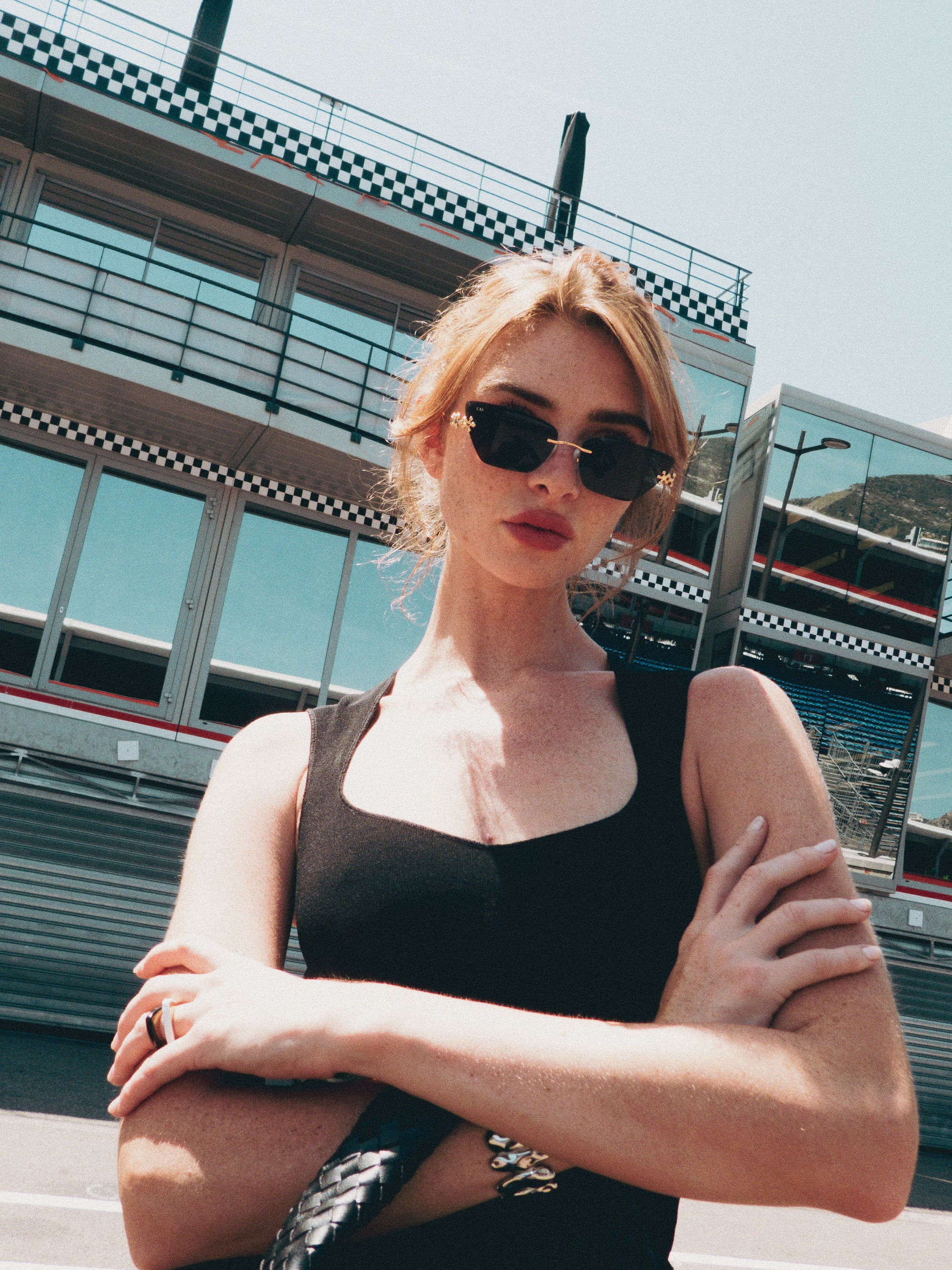 A woman in For Art's Sake® Florist floral sunglasses and a black sleeveless top stands with arms crossed before a modern racing stadium with glass windows and checkered details.