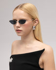 A woman with straight blonde hair wears For Art's Sake® Lake cat-eye sunglasses with sculpted temples, large rectangular earrings, and an off-shoulder black dress featuring a jeweled brooch, posing before a plain light gray background.
