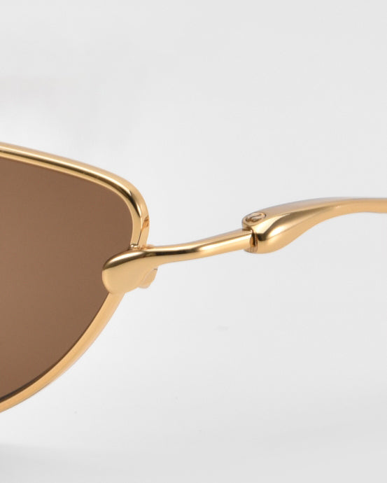 Close-up of the For Art’s Sake® Lake cat-eye sunglasses in gold metal with brown tinted lenses, highlighting the corner of the lens and sculpted temples against a plain light background.