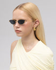 A woman with straight blonde hair wears For Art’s Sake® Lake cat-eye sunglasses, gold earrings, and a pale yellow sleeveless top against a plain light background.