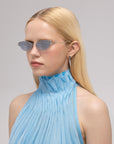 A woman with straight blonde hair wears For Art's Sake® Lake oval-shaped blue sunglasses with sculpted temples and a sleeveless, high-neck pleated blue top. She has a neutral expression, small hoop earring, and stands against a plain gray background.