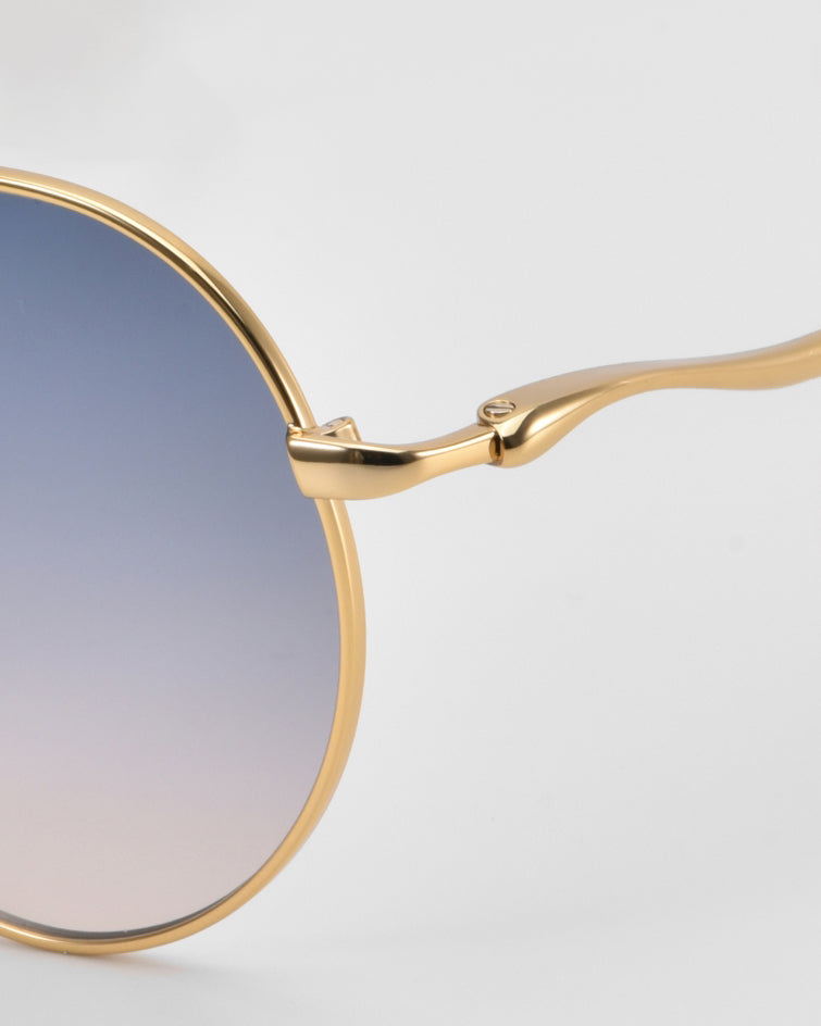 Close-up of For Art's Sake® Lines: oversized aviator sunglasses with a gold frame and gradient blue-to-clear lenses. Sculpted temples and hinge detail stand out against a plain light gray background, showcasing the brand’s signature style.