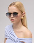 A woman with straight blonde hair wears For Art's Sake® Lines oversized aviator sunglasses with sculpted temples, gold square earrings, and a light blue off-the-shoulder top, looking at the camera against a plain background.
