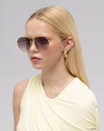 A woman with long blonde hair wears oversized Lines aviator sunglasses by For Art’s Sake®, gold geometric earrings, and a light yellow sleeveless top against a plain light gray background.