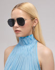 A woman with long blonde hair wears oversized aviator sunglasses—Lines by For Art's Sake®—featuring sculpted temples, along with a sleeveless pleated light blue high-collar top and geometric silver earrings, set against a plain light background.