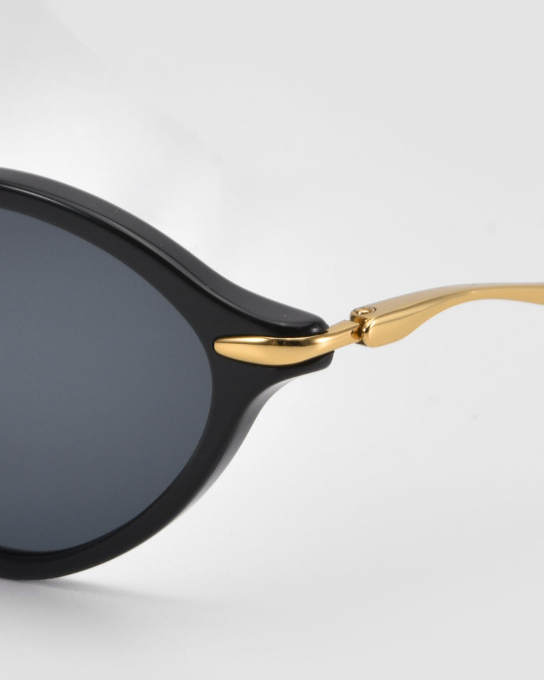 Close-up of For Art's Sake® Totto sunglasses featuring a black acetate and metal frame, sculpted temples, and round dark lenses against a light background.