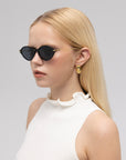 A woman with long blonde hair wears round black For Art's Sake® Totto sunglasses with sculpted temples and gold earrings, paired with a white sleeveless top featuring a ruffled collar, standing against a plain light background.
