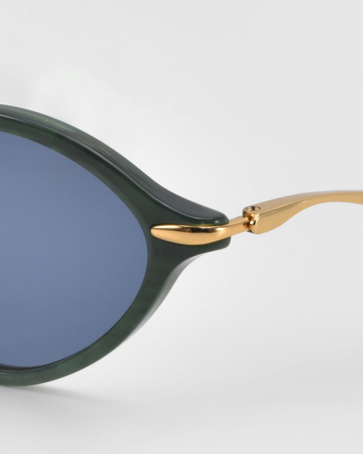 Close-up of For Art's Sake® Totto sunglasses featuring a green acetate and metal frame, blue-tinted lenses, and gold sculpted temples on a plain light gray background.