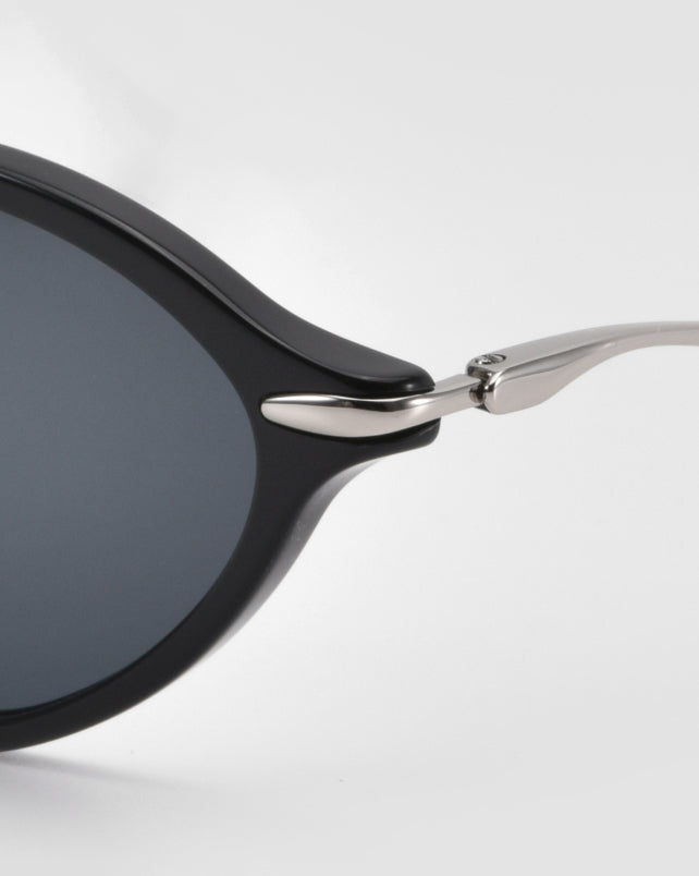 Close-up of the hinge and frame corner of For Art's Sake® Totto sunglasses with sleek acetate and metal construction, dark lenses, and a silver arm against a light gray background.