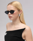 A woman with straight blonde hair wears black For Art's Sake® Totto sunglasses with sculpted temples, square earrings, and a strapless black dress adorned with a jeweled brooch, standing against a plain light gray background.