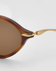 Close-up of For Art's Sake® Totto sunglasses with brown-tinted lenses, a brown frame, sculpted acetate and metal temples, plus gold metallic hinge and temple arm against a plain light background.