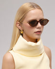 A woman with straight blonde hair wears For Art's Sake® Totto oval brown sunglasses with sculpted acetate and metal temples, gold earrings, and a sleeveless pale yellow top with a structured collar against a neutral background.