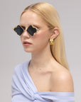 A woman with long blonde hair models For Art's Sake® Leaf I sculptural glasses, gold geometric earrings, and a light blue off-shoulder top against a plain light gray background.