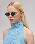 A woman with long blonde hair models For Art's Sake® Leaf I sculptural sunglasses and a light blue pleated sleeveless top with a high ruffled collar, set against a neutral background.