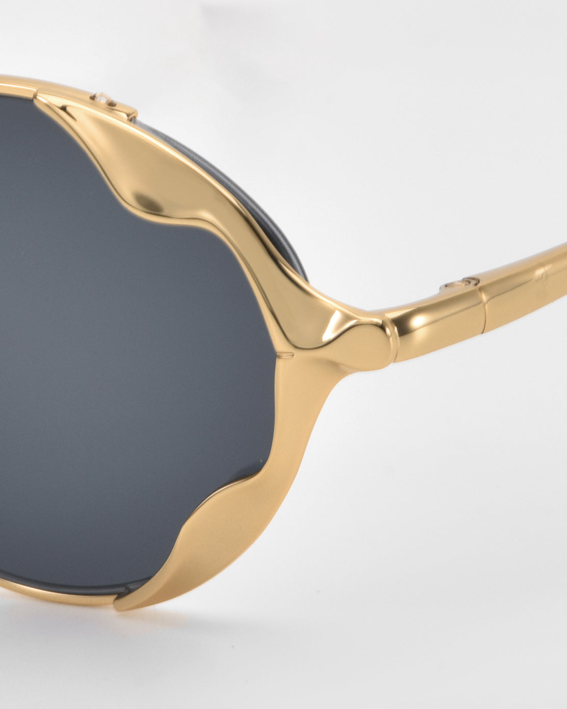 Close-up of For Art's Sake® Leaf II sunglasses featuring dark lenses and a distinctive wavy gold metal frame, creating bold statement eyewear against a plain light background.
