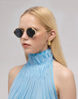 A woman with long blonde hair wears For Art's Sake® Leaf II sculptural sunglasses with round, gold-rimmed black lenses, gold earrings, and a sleeveless light blue pleated top against a neutral background.