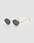 The Leaf II round sunglasses by For Art's Sake® feature gold metal frames and dark lenses, shown on a light gray background.