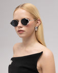 A woman with straight blonde hair wears For Art's Sake® Leaf II sunglasses, geometric earrings, and an off-shoulder black dress with a brooch. The background is plain light gray.