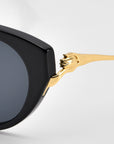 Close-up of For Art’s Sake® Tulip I sunglasses in bold acetate with dark lenses, featuring a gold temple shaped like a panther’s head and paw gripping the frame. Set against a plain light gray background for an elegant look.