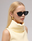 A woman with straight blonde hair wears For Art's Sake® Tulip I bold cat-eye acetate sunglasses, gold earrings, and a pale yellow high-neck sleeveless top, gazing slightly to the side against a plain background.