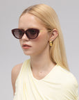 A woman with long blonde hair wears For Art's Sake® Tulip I bold acetate sunglasses, gold geometric earrings, and a sleeveless light yellow top while looking off to the side against a plain gray background.