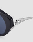 Close-up of For Art's Sake® Tulip I sunglasses with bold acetate frames and a sculptural silver temple shaped like a hand gripping the hinge. Contrasting dark lenses are set against a light grey background.