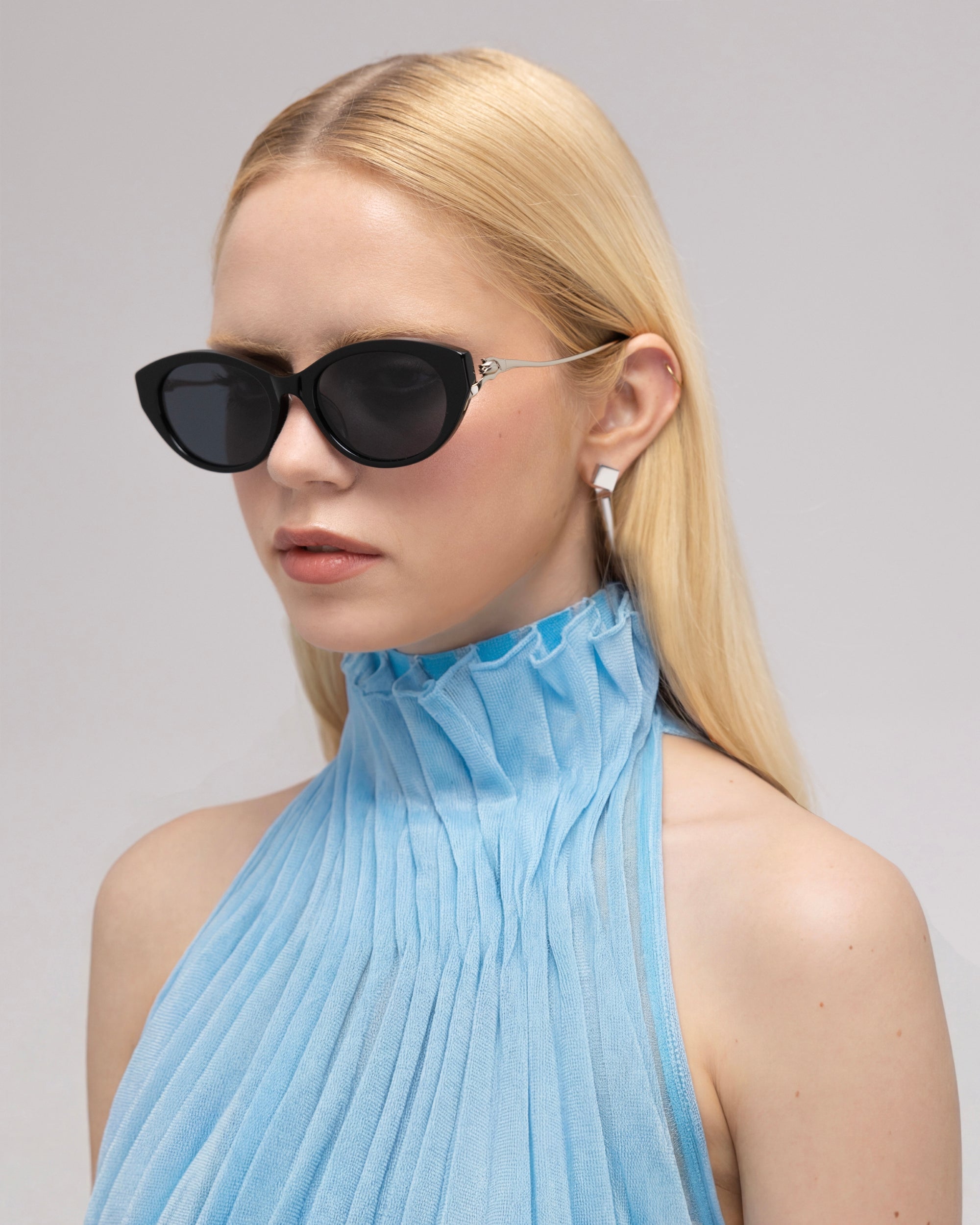 A woman with long blonde hair models For Art's Sake® Tulip I bold acetate sunglasses and silver earrings, paired with a light blue pleated sleeveless top, against a plain light background.