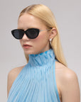 A woman with long blonde hair models For Art's Sake® Tulip I bold acetate sunglasses and silver earrings, paired with a light blue pleated sleeveless top, against a plain light background.