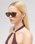 A woman with straight blonde hair models For Art's Sake® Tulip I brown sunglasses with an acetate front, gold earrings, and a mauve sleeveless top with a gold accent, against a plain light background.