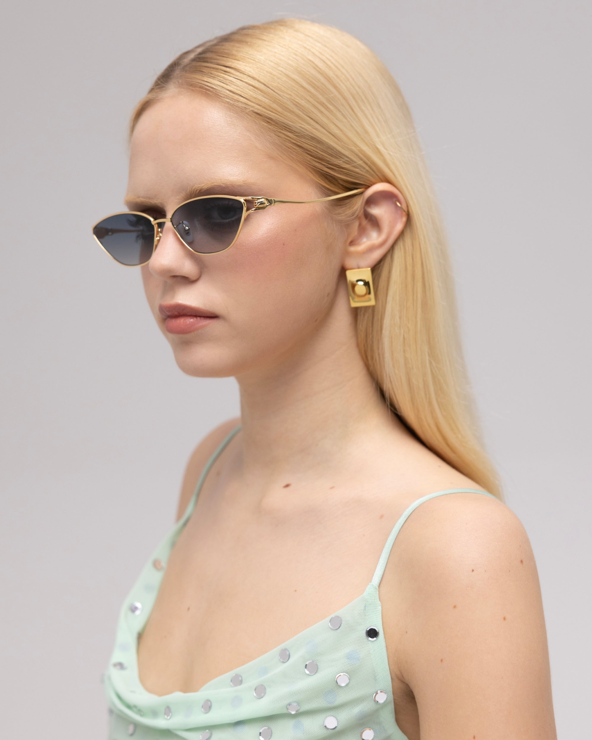 A woman with straight blonde hair wears For Art's Sake® Tulip II black cat-eye sunglasses with a bold acetate front, large gold earrings, and a light green polka-dot dress, gazing to the side against a neutral background.