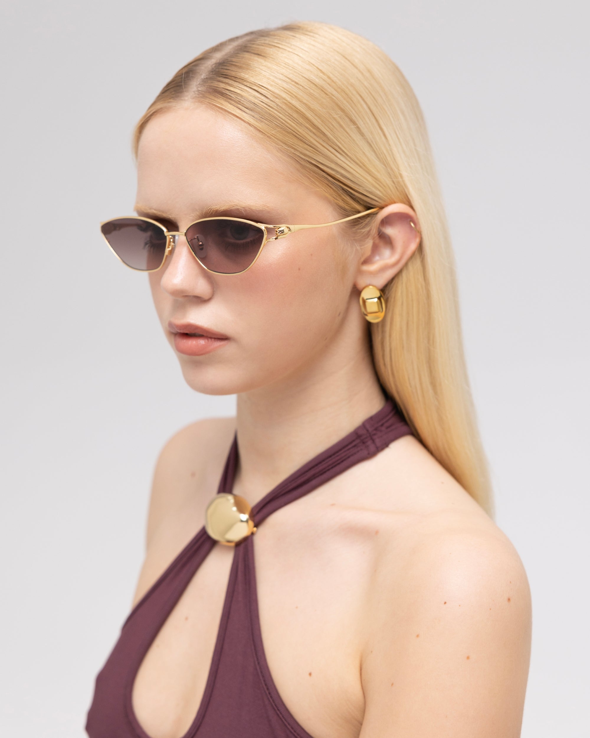 A woman with straight blonde hair wears For Art's Sake® Tulip II bold acetate sunglasses, gold geometric earrings, and a dark halter top with a gold accent, posing against a plain light background.