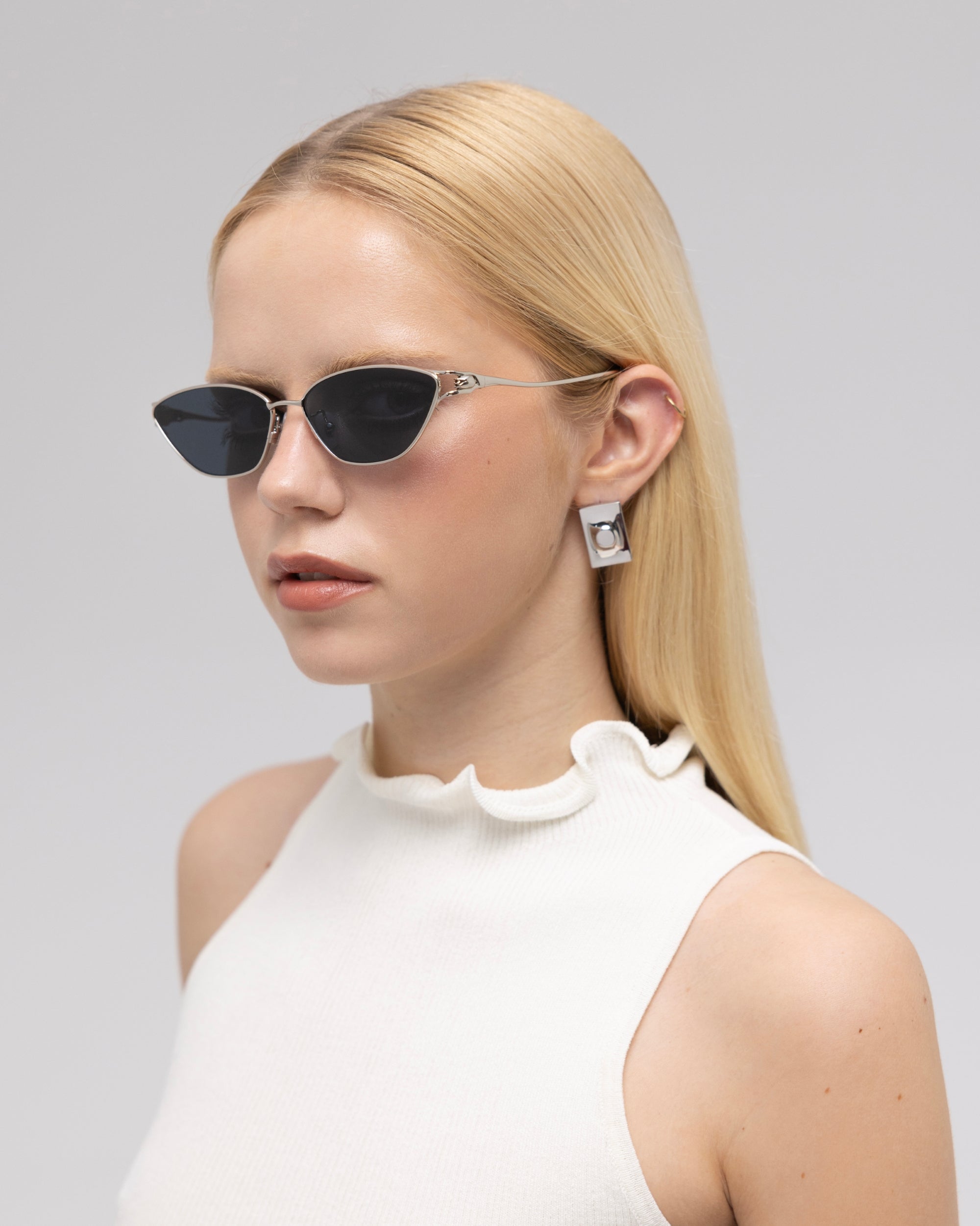 A woman with straight blonde hair models For Art's Sake® Tulip II bold acetate sunglasses, paired with rectangular silver earrings and a sleeveless white ruffled top, posing before a light grey backdrop.