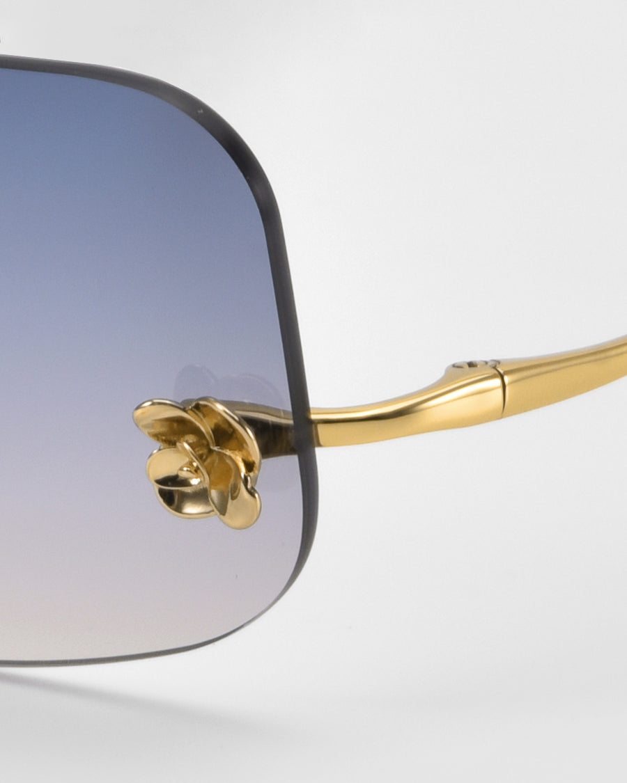 Close-up of For Art's Sake® Magnolia sunglasses, showcasing a gold frame, gradient blue-to-clear lenses, and a signature decorative gold flower detail at the hinge.