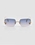 The Magnolia by For Art’s Sake® are sculptural, frameless sunglasses with gradient blue lenses, a gold nose bridge, and thin gold arms, shown against a plain light gray background.