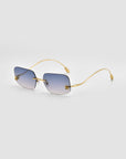 The Magnolia by For Art's Sake® are square, frameless sunglasses with gradient blue lenses and slim gold metal temples, shown on a light gray background.