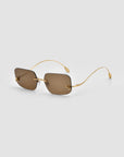 The Magnolia sunglasses by For Art's Sake® feature sculptural, rimless rectangular brown-tinted lenses with slim gold metal arms and nose bridge, shown on a light gray background.