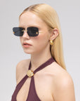 A woman with straight blonde hair models For Art's Sake® Magnolia frameless sunglasses, gold earrings, and a sleeveless maroon top with a gold circular clasp at the neckline, set against a plain light background.