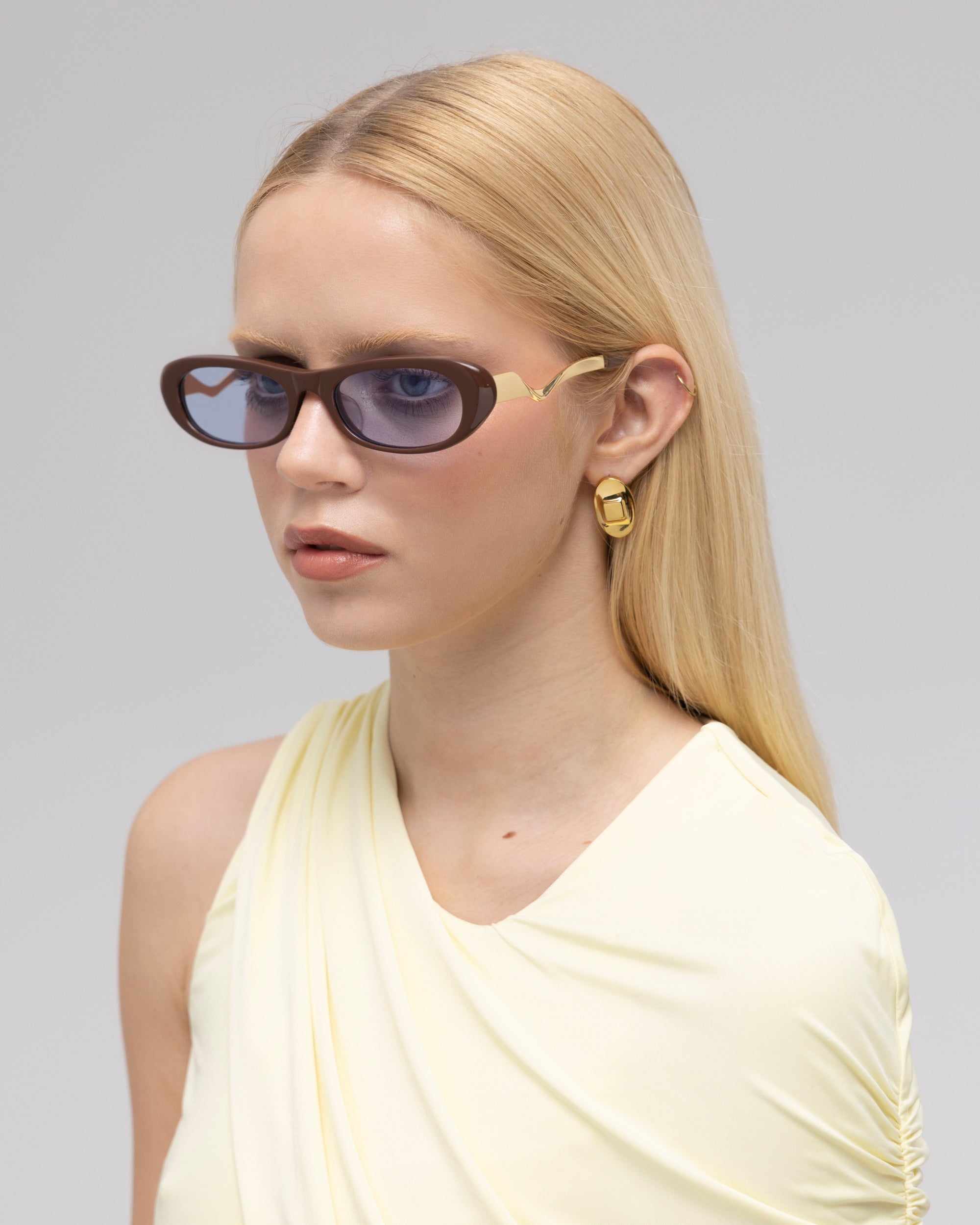 A woman with long, straight blonde hair models For Art's Sake® Abstraction I sculptural metal sunglasses and gold earrings, wearing a pale yellow sleeveless top, facing slightly right against a plain light gray background.