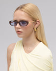 A woman with long, straight blonde hair models For Art's Sake® Abstraction I sculptural metal sunglasses and gold earrings, wearing a pale yellow sleeveless top, facing slightly right against a plain light gray background.