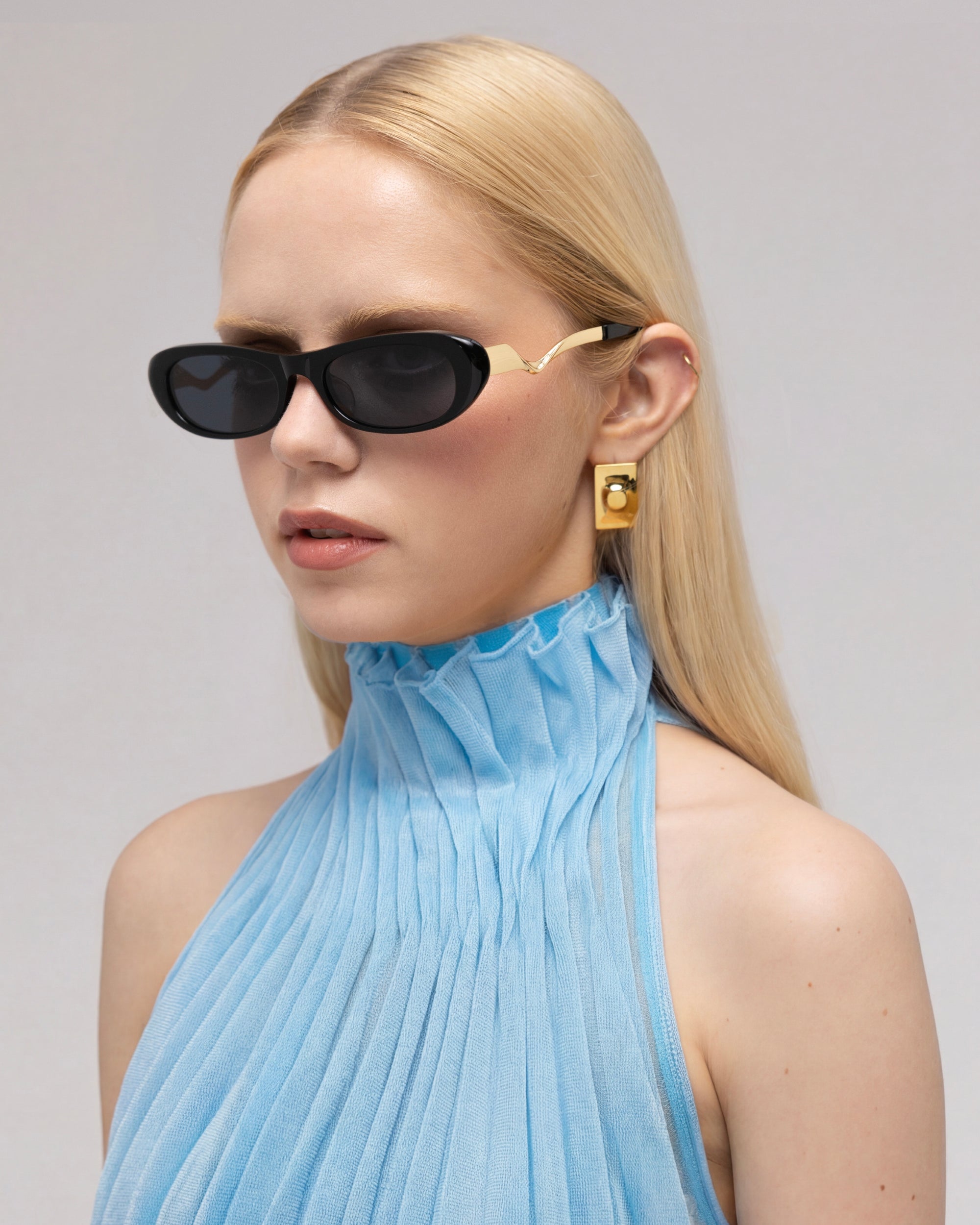 A woman with straight blonde hair models For Art’s Sake® Abstraction I bold temple sunglasses, gold earrings, and a light blue pleated sleeveless top against a simple, light background.